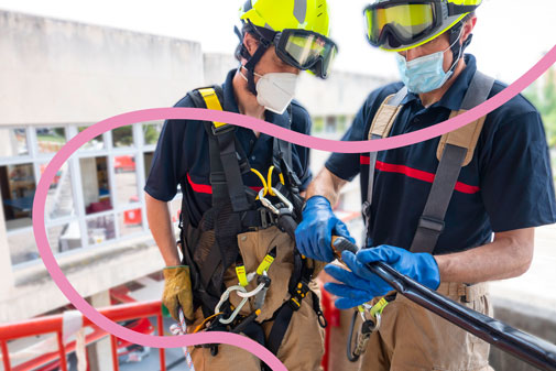 Dos homes amb casc i arnès revisen mosquetons d'un equip de protecció anticaigudes.